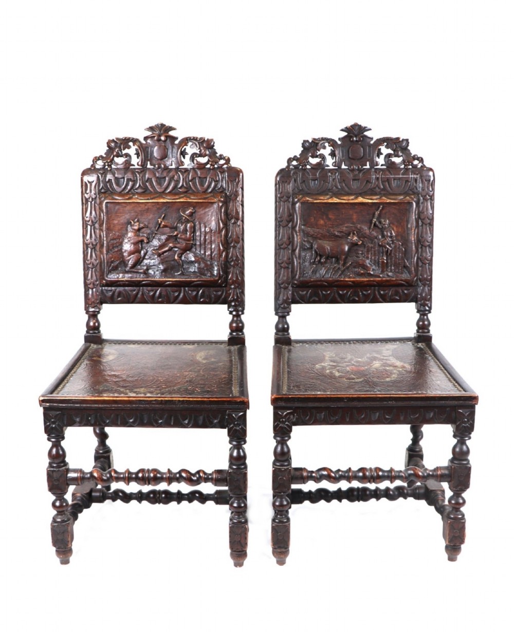 A pair of dark carved wooden side chairs featuring high-relief back panels depicting figurative scenes, leather seats, and turned stretchers.