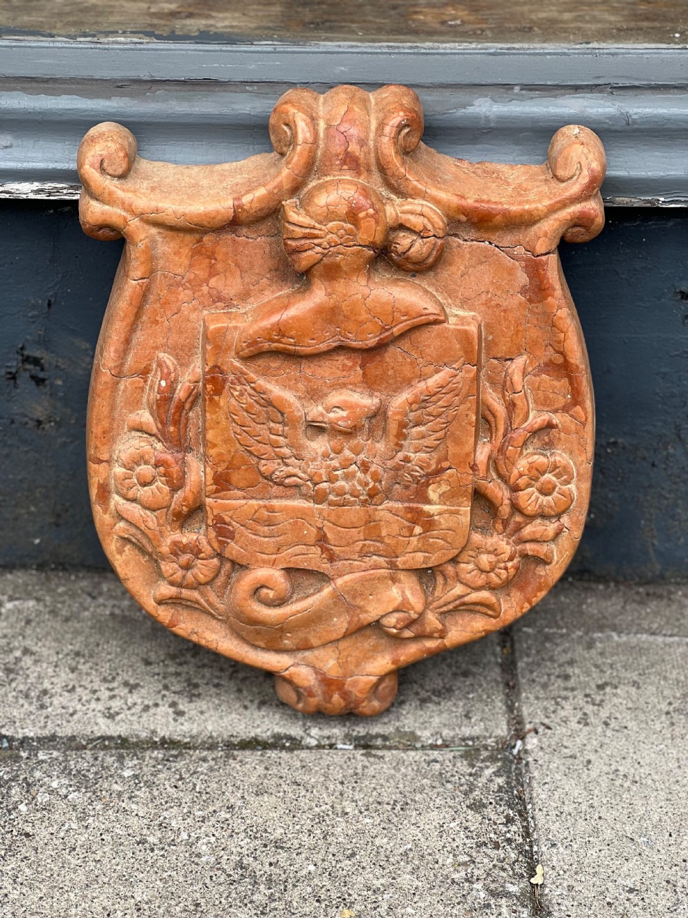 A carved terracotta-red marble armorial crest featuring a central shield with an eagle motif framed by carved flowers and scrollwork.