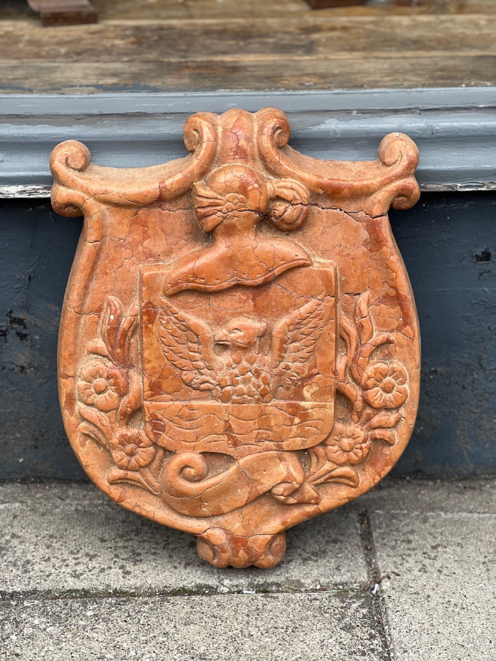 A carved terracotta-red marble armorial crest featuring a central shield with an eagle motif framed by carved flowers and scrollwork. View II