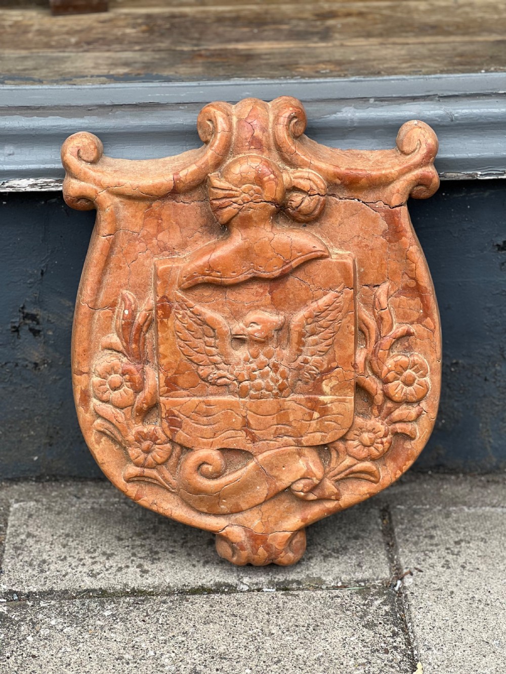 A carved terracotta-red marble armorial crest featuring a central shield with an eagle motif framed by carved flowers and scrollwork. View III