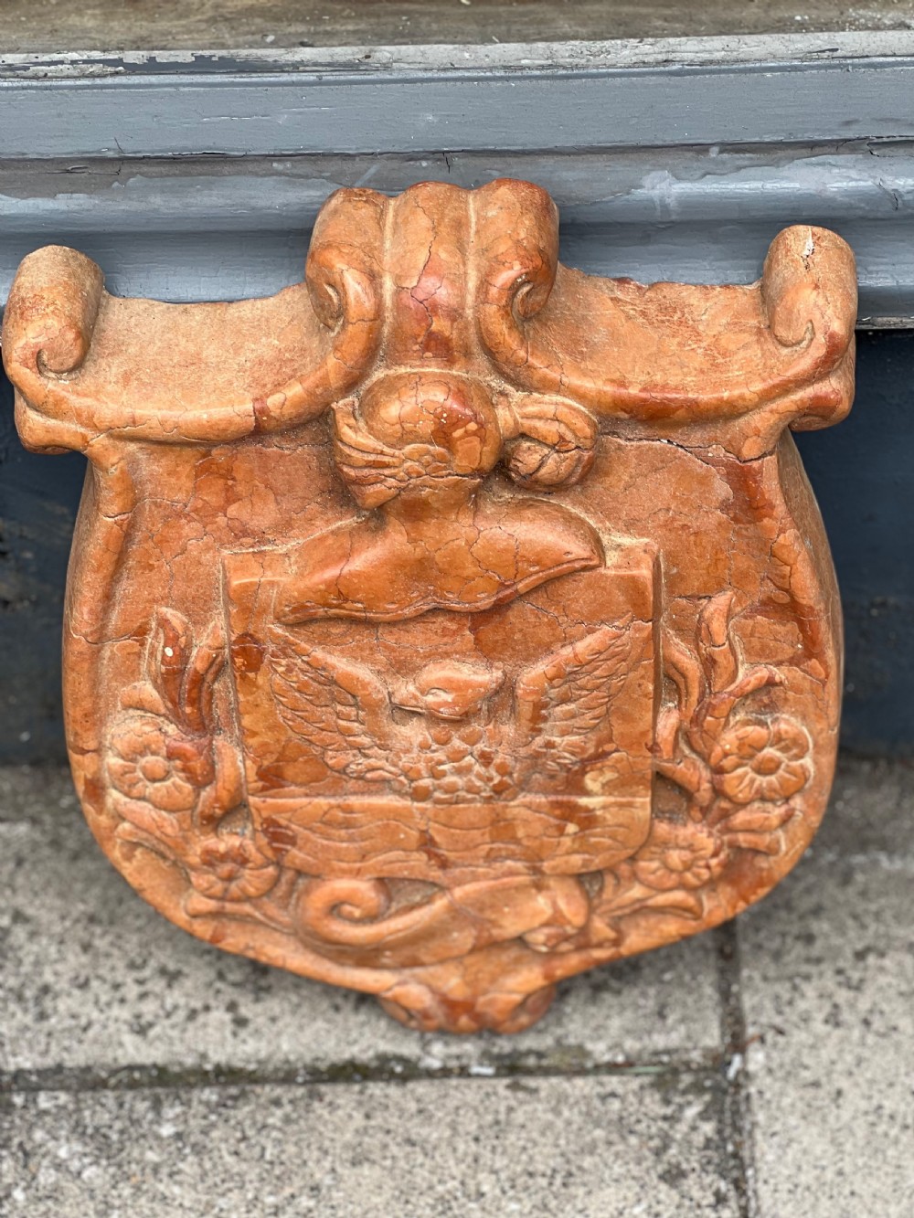 A carved terracotta-red marble armorial crest featuring a central shield with an eagle motif framed by carved flowers and scrollwork. View V