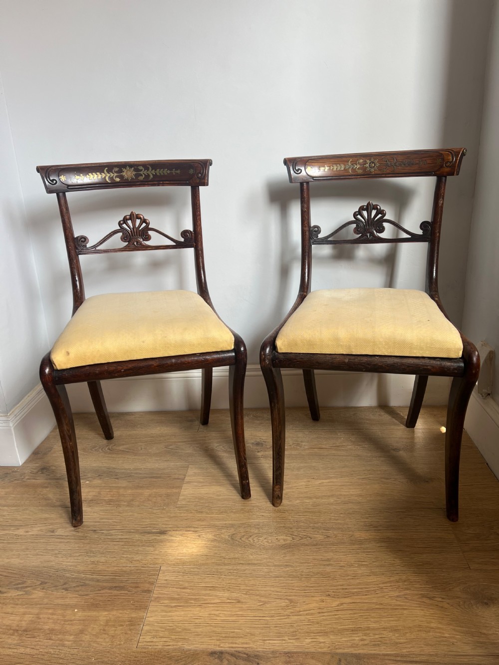 A pair of Regency faux rosewood side chairs featuring brass inlaid crest rails, carved anthemion splats, sabre legs, and pale yellow upholstered seats.
