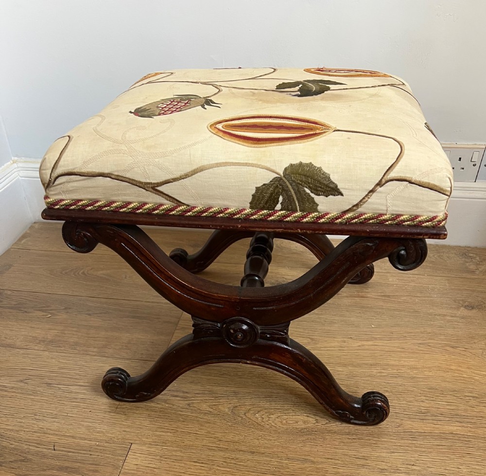 A 19th-century mahogany X-frame stool featuring carved scroll legs, a turned central stretcher, and a cream floral embroidered upholstered cushion.