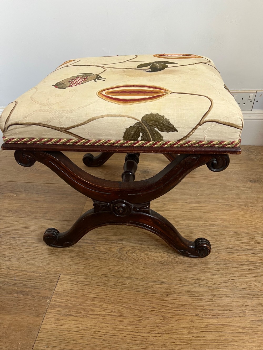 A 19th-century mahogany X-frame stool featuring carved scroll legs, a turned central stretcher, and a cream floral embroidered upholstered cushion. View V