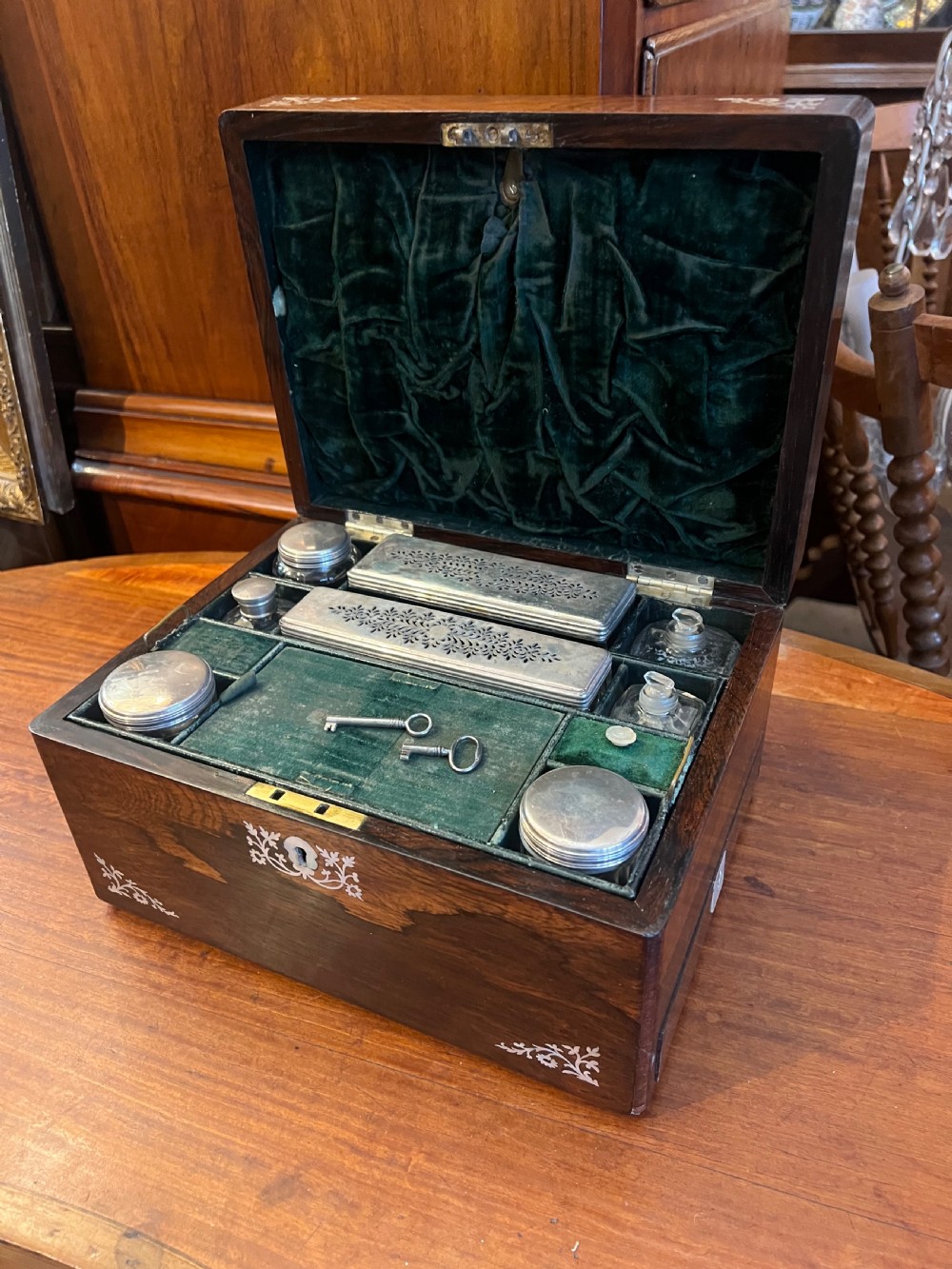 An open Victorian rosewood workbox featuring floral mother of pearl inlay, fitted green velvet interior, silver-toned vanity jars, glass bottles, and two small keys.