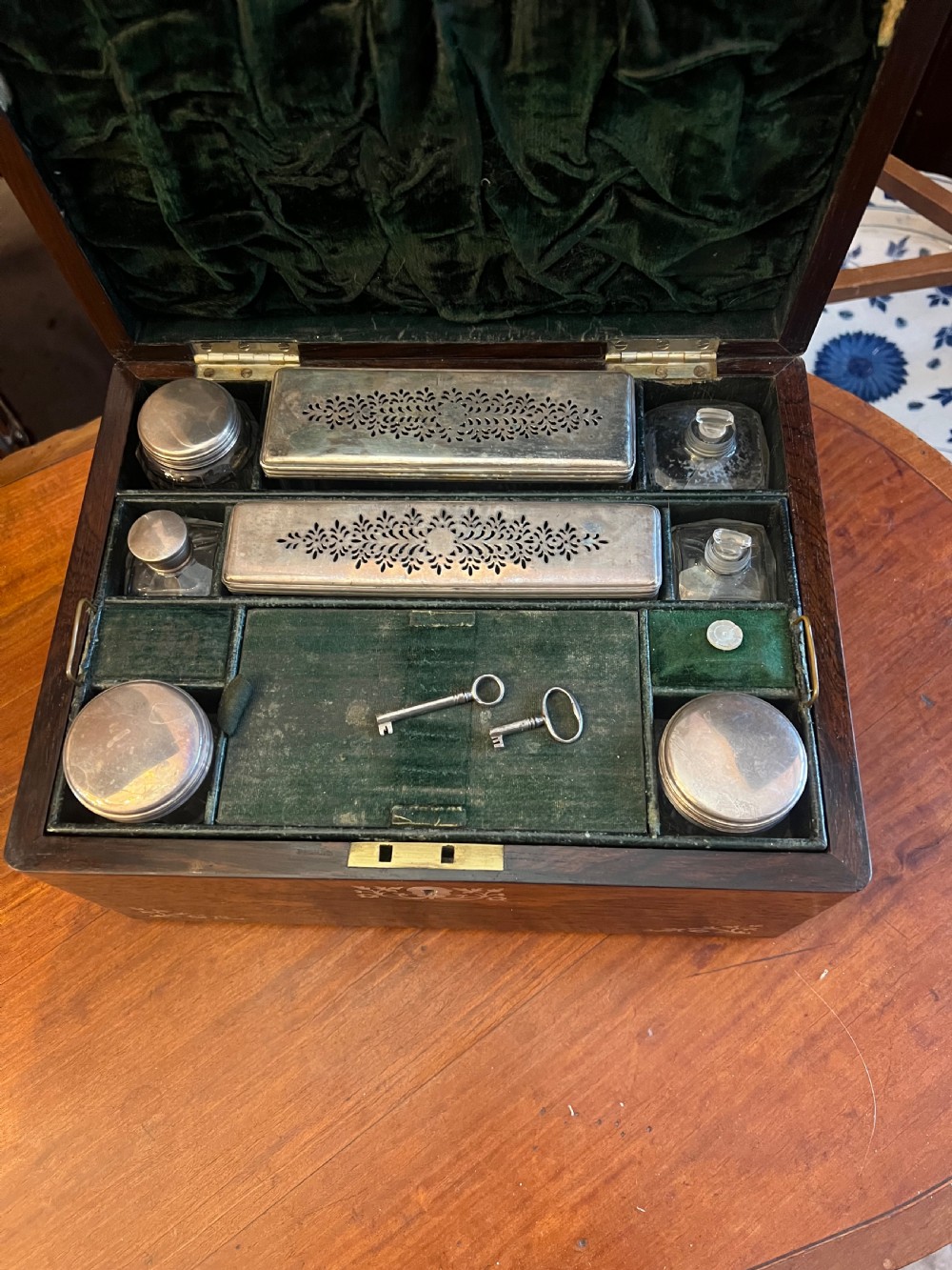 An open Victorian rosewood workbox featuring floral mother of pearl inlay, fitted green velvet interior, silver-toned vanity jars, glass bottles, and two small keys. View XVI