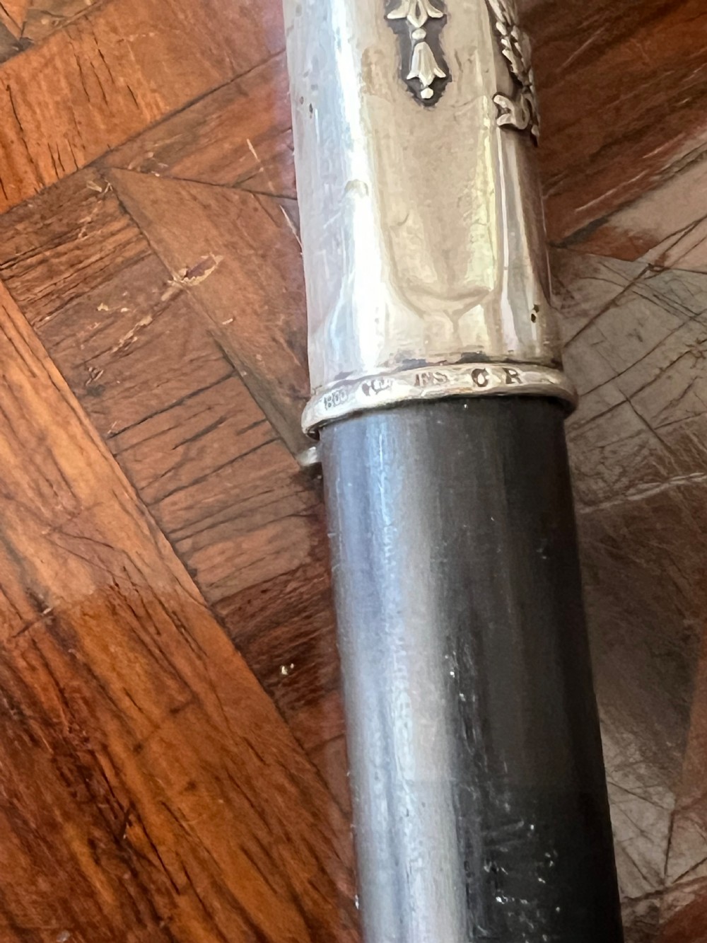 An antique walking stick featuring an embossed silver crook handle attached to a polished black wooden shaft. View V