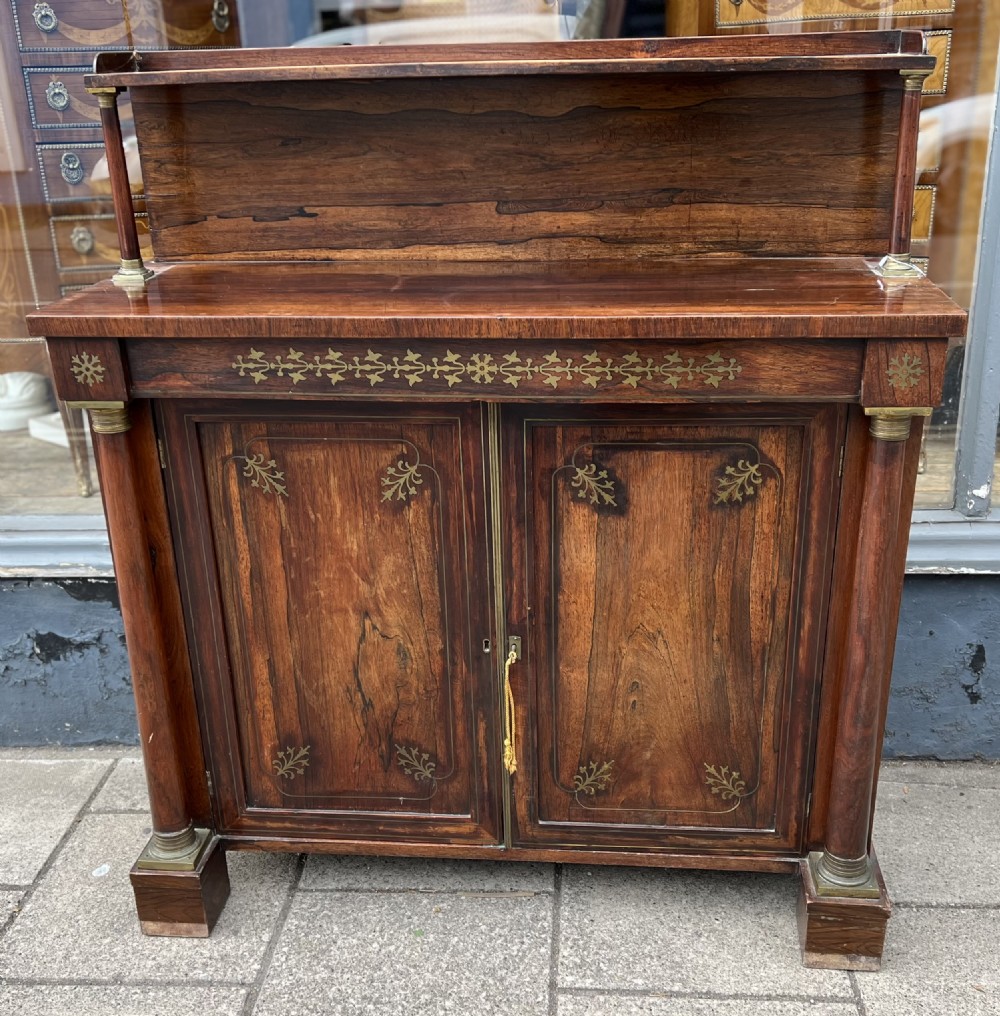 An antique Regency dark rosewood side cabinet featuring brass floral inlay, twin panelled doors, side columns with brass capitals, and an upper shelf resting on turned brass pillars.