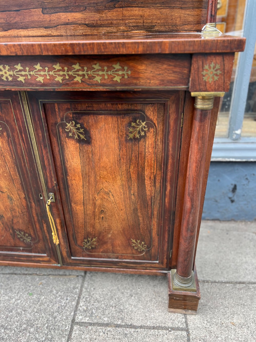 An antique Regency dark rosewood side cabinet featuring brass floral inlay, twin panelled doors, side columns with brass capitals, and an upper shelf resting on turned brass pillars. View II