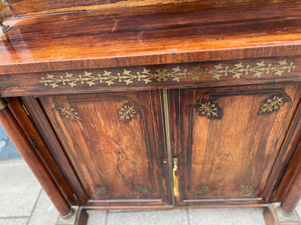 An antique Regency dark rosewood side cabinet featuring brass floral inlay, twin panelled doors, side columns with brass capitals, and an upper shelf resting on turned brass pillars. View III
