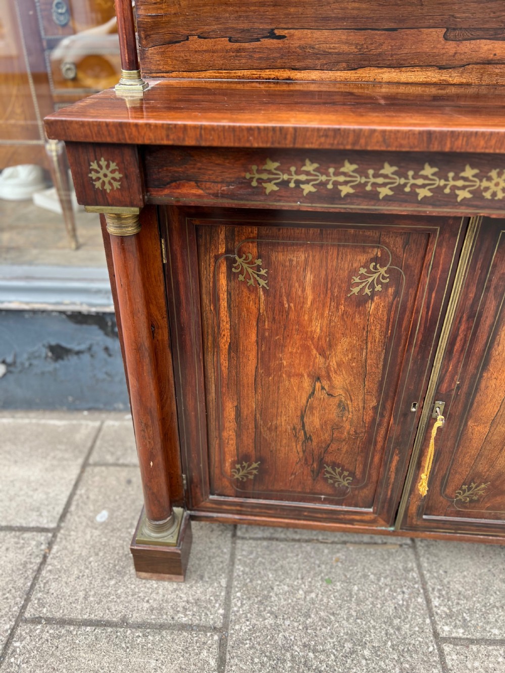 An antique Regency dark rosewood side cabinet featuring brass floral inlay, twin panelled doors, side columns with brass capitals, and an upper shelf resting on turned brass pillars. View V