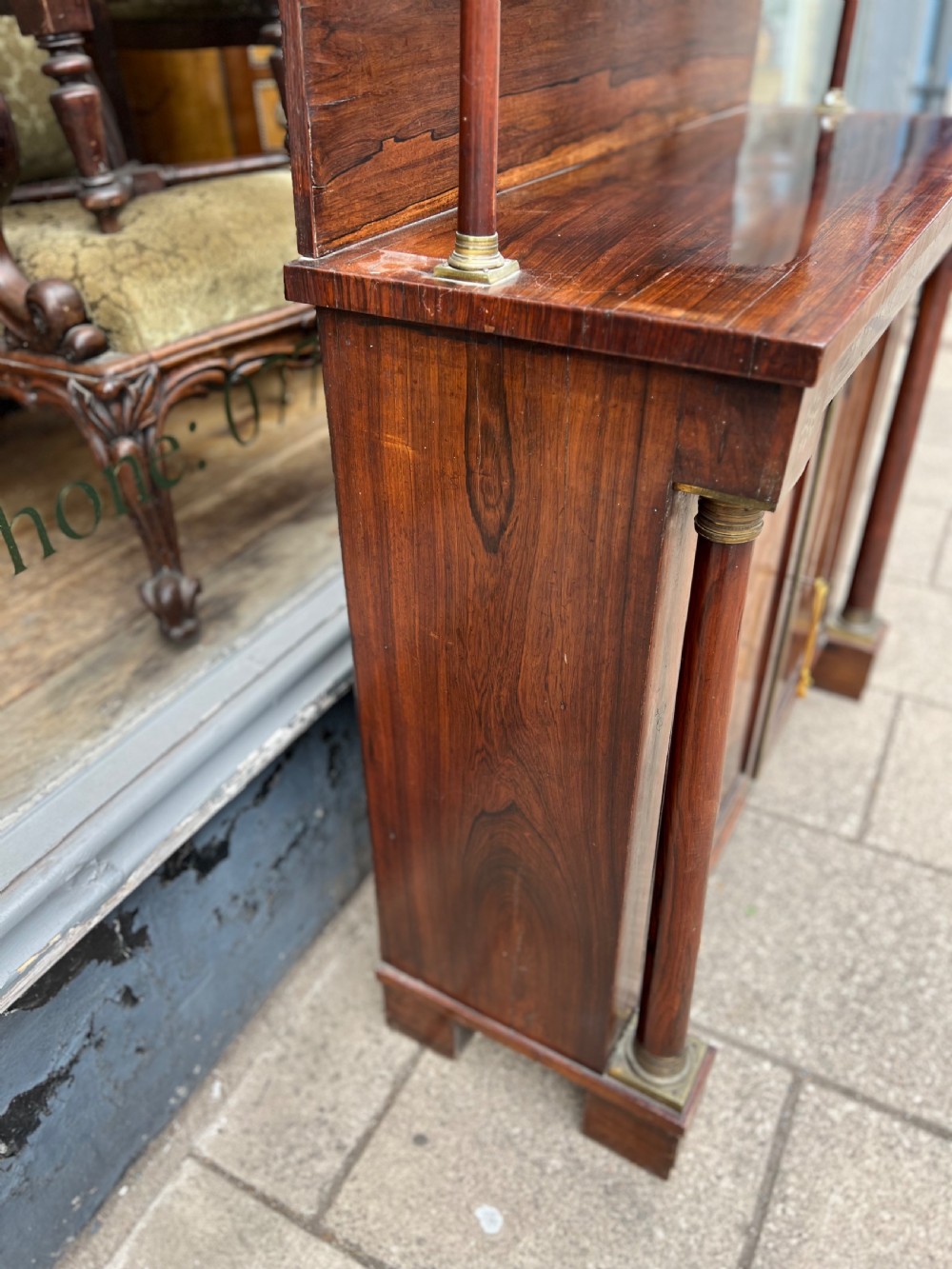 An antique Regency dark rosewood side cabinet featuring brass floral inlay, twin panelled doors, side columns with brass capitals, and an upper shelf resting on turned brass pillars. View VI