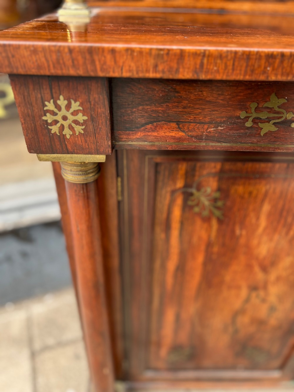 An antique Regency dark rosewood side cabinet featuring brass floral inlay, twin panelled doors, side columns with brass capitals, and an upper shelf resting on turned brass pillars. View XIII