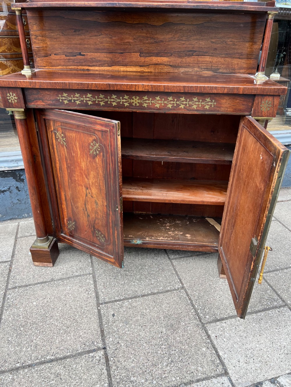 An antique Regency dark rosewood side cabinet featuring brass floral inlay, twin panelled doors, side columns with brass capitals, and an upper shelf resting on turned brass pillars. View XV