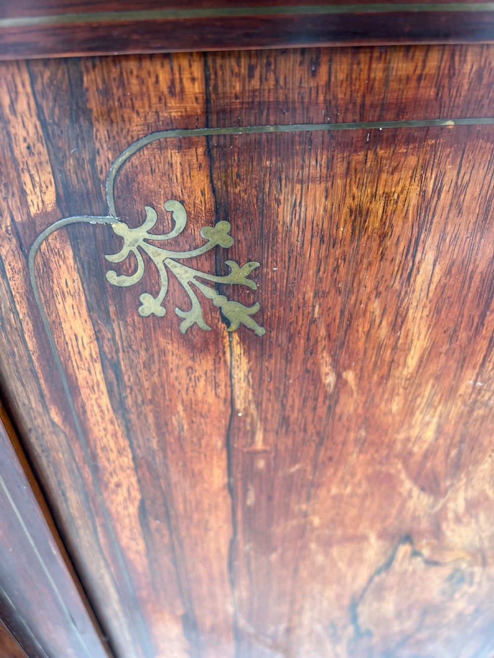 An antique Regency dark rosewood side cabinet featuring brass floral inlay, twin panelled doors, side columns with brass capitals, and an upper shelf resting on turned brass pillars. View XVI