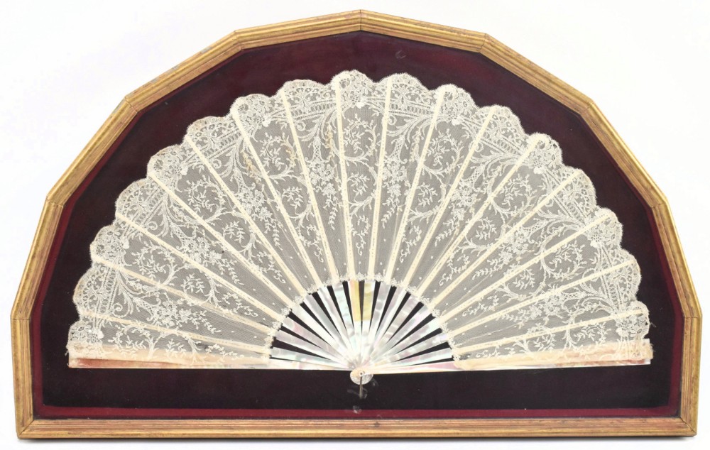 A 19th-century lace fan with iridescent mother of pearl sticks mounted against burgundy velvet inside an arched gilt frame.