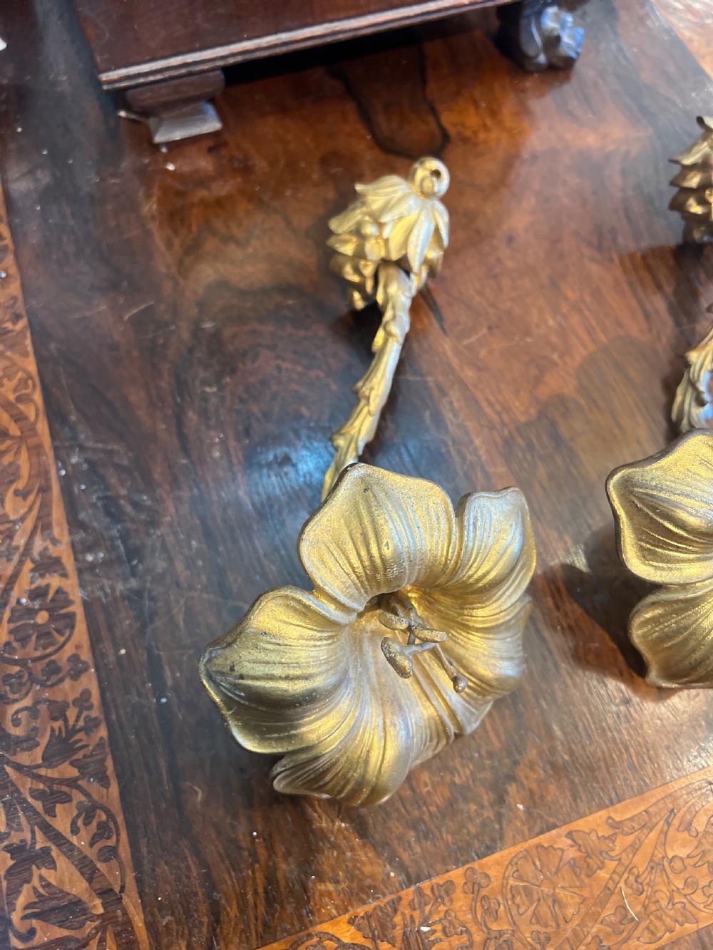 A pair of cast gilt brass curtain tiebacks featuring a stylised flower head design and textured stem. View II