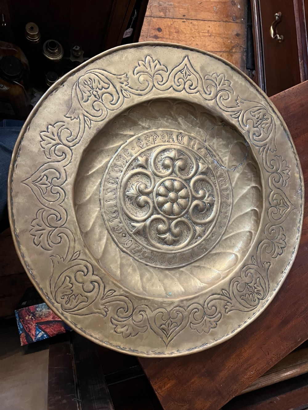 A circular 18th-century brass alms dish featuring an embossed central floral boss, a ring of gothic lettering, and a foliate patterned rim. View II