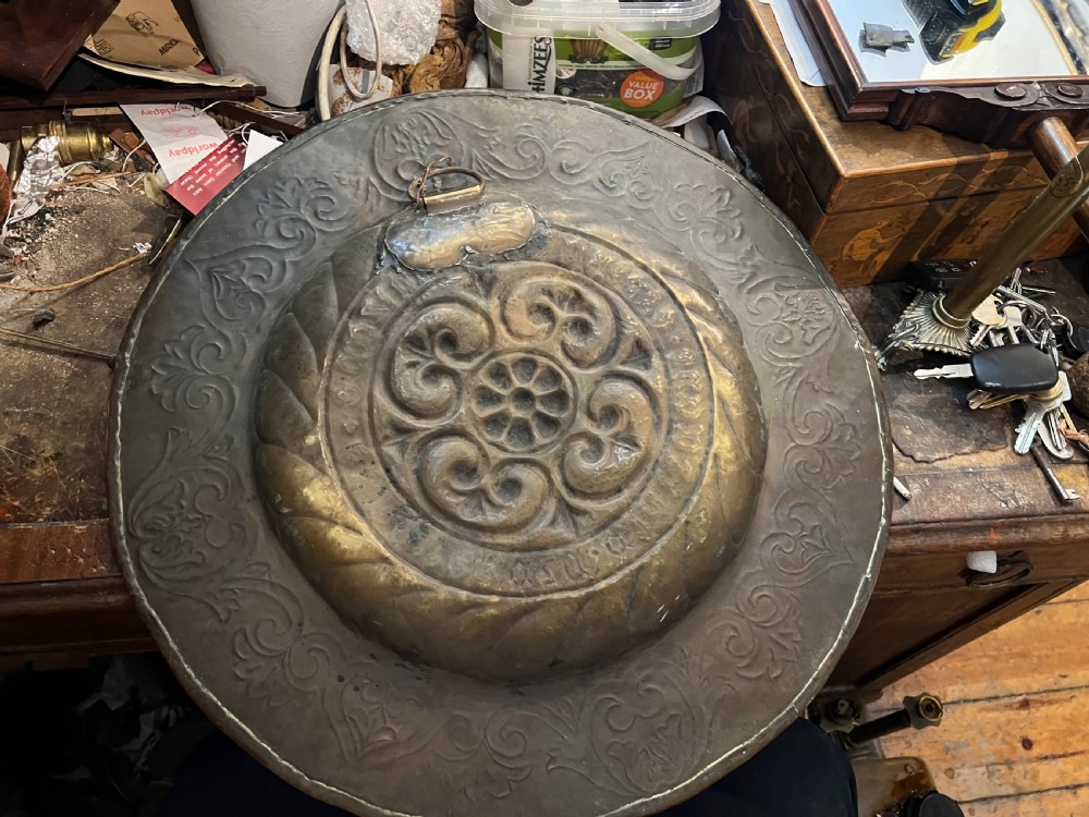 A circular 18th-century brass alms dish featuring an embossed central floral boss, a ring of gothic lettering, and a foliate patterned rim. View V