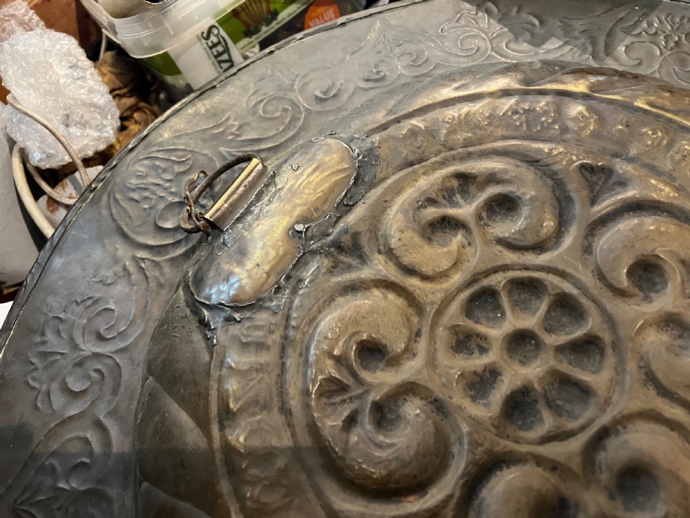 A circular 18th-century brass alms dish featuring an embossed central floral boss, a ring of gothic lettering, and a foliate patterned rim. View VI