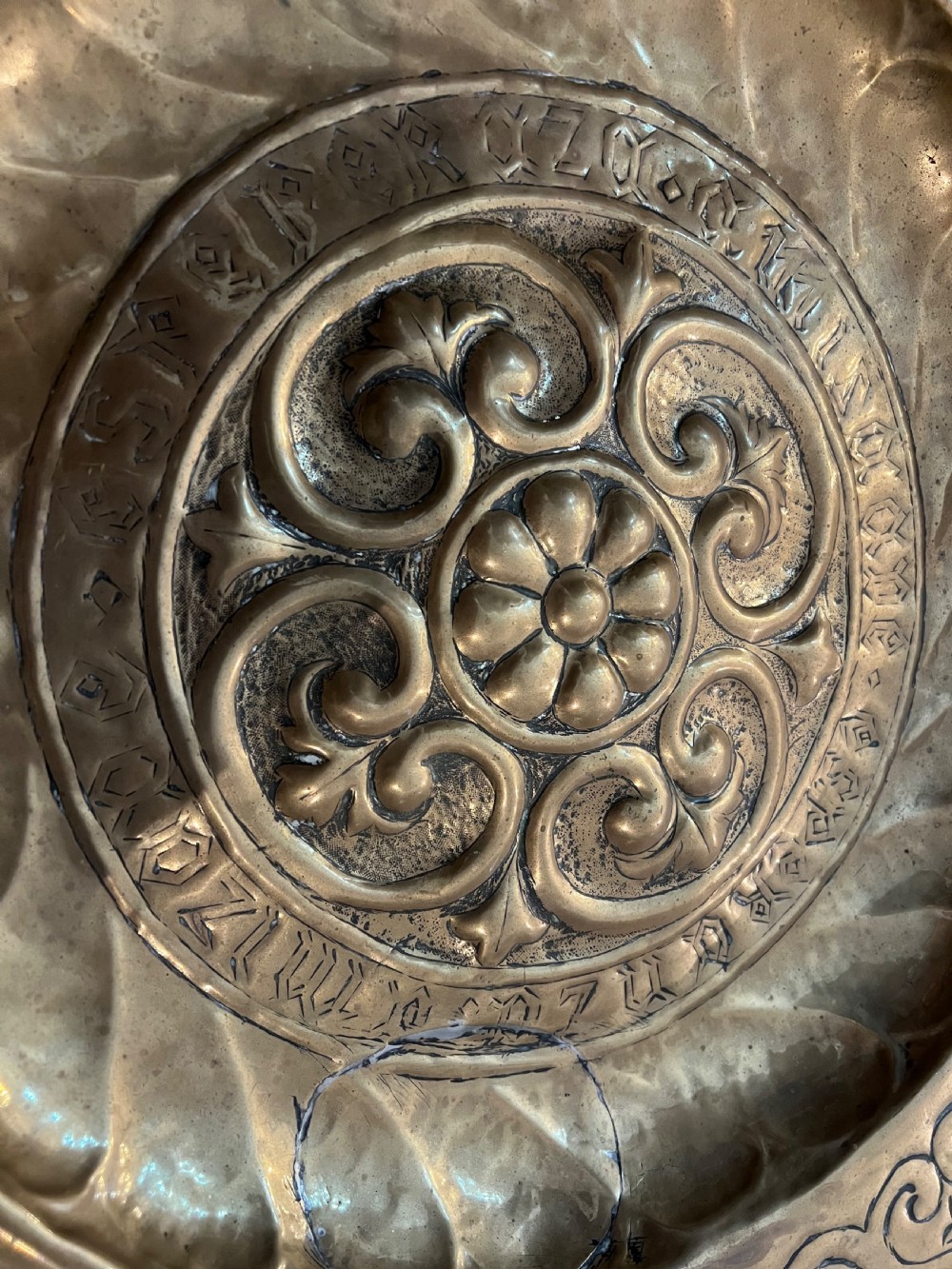 A circular 18th-century brass alms dish featuring an embossed central floral boss, a ring of gothic lettering, and a foliate patterned rim. View IX