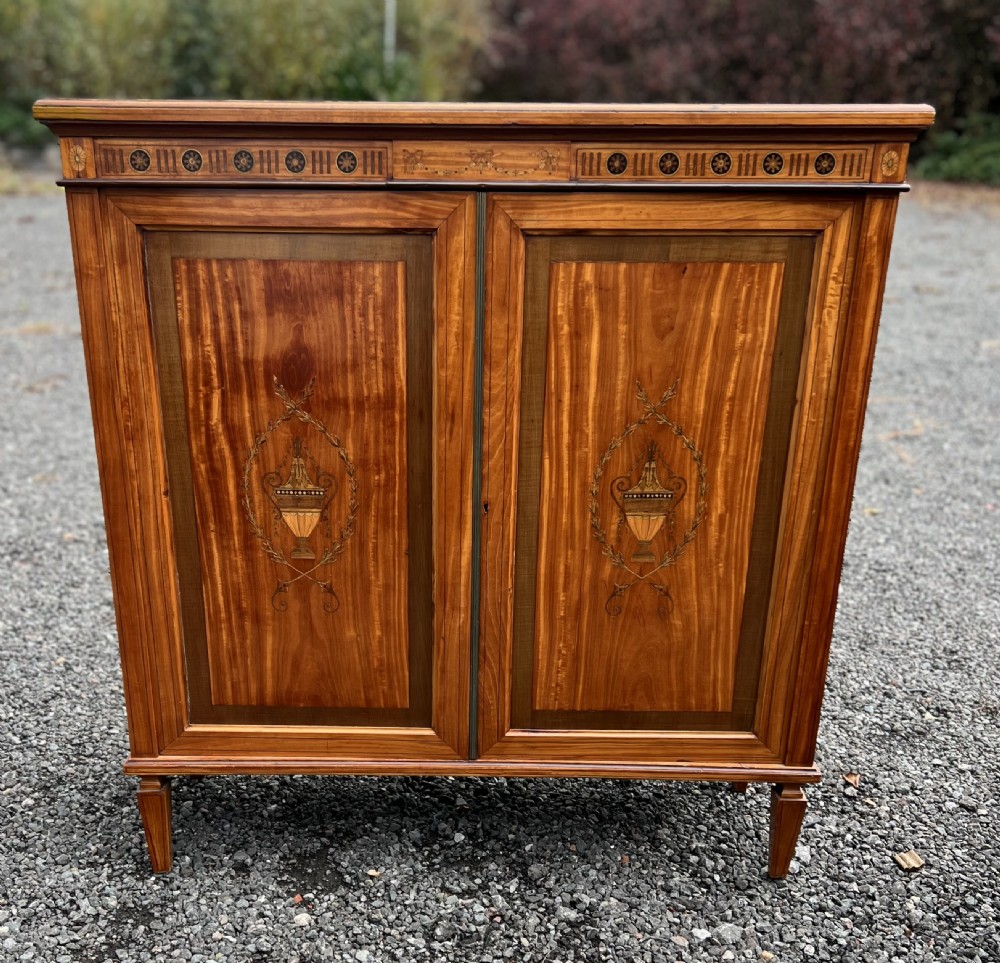 A satinwood side cabinet featuring two cupboard doors with central marquetry urn inlays, a decorated top frieze, and square tapered feet. View II