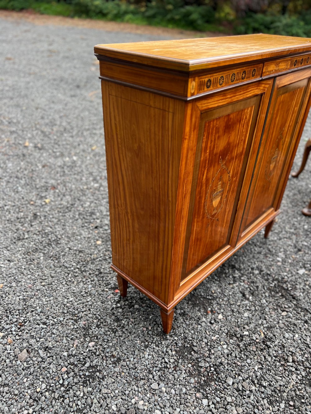 A satinwood side cabinet featuring two cupboard doors with central marquetry urn inlays, a decorated top frieze, and square tapered feet. View III