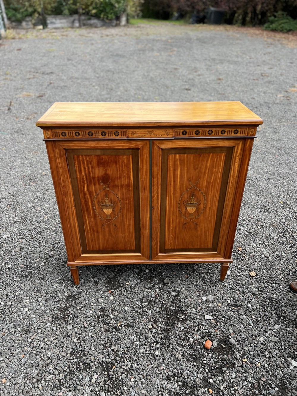 A satinwood side cabinet featuring two cupboard doors with central marquetry urn inlays, a decorated top frieze, and square tapered feet. View VII