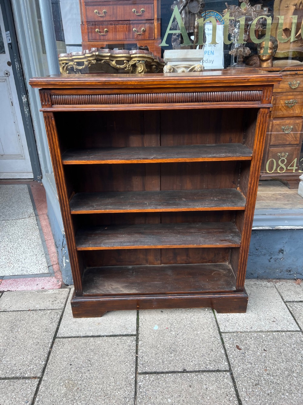 An antique open Edwardian oak bookcase featuring four storage levels, fluted side pilasters, and a carved frieze above a solid plinth base.