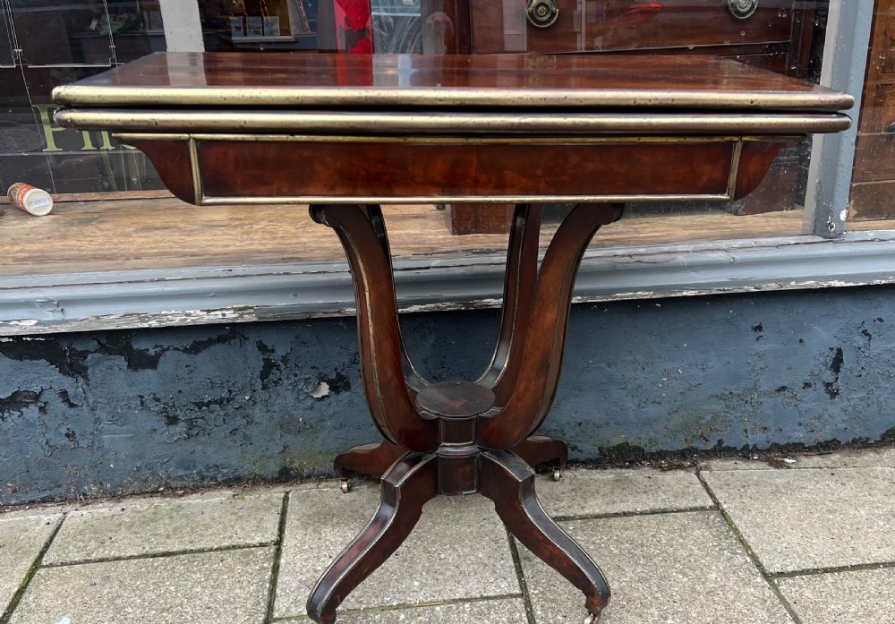 A Regency mahogany card table with brass edging, fold-over top, and quadform pedestal base on castors.