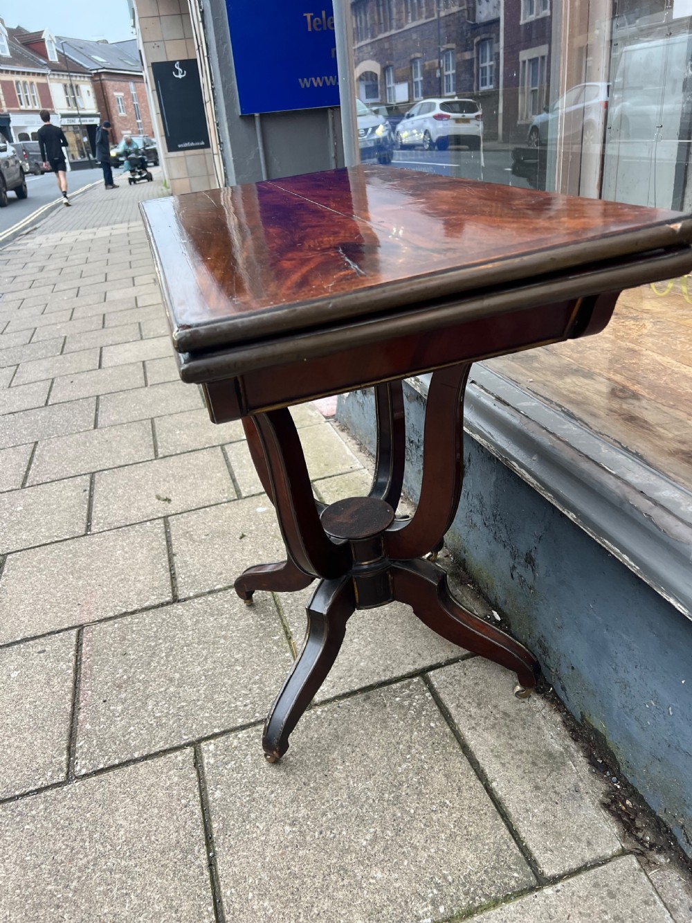 A Regency mahogany card table with brass edging, fold-over top, and quadform pedestal base on castors. View II