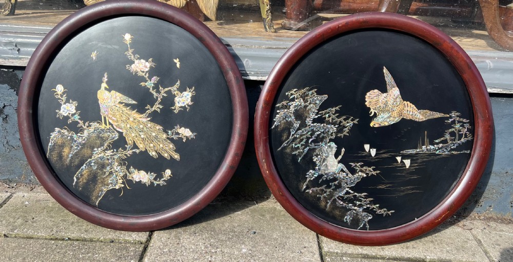 A pair of circular black lacquered wall plaques set in dark wood frames, decorated with mother-of-pearl and bone inlays depicting a peacock amongst foliage and a bird in flight over water.