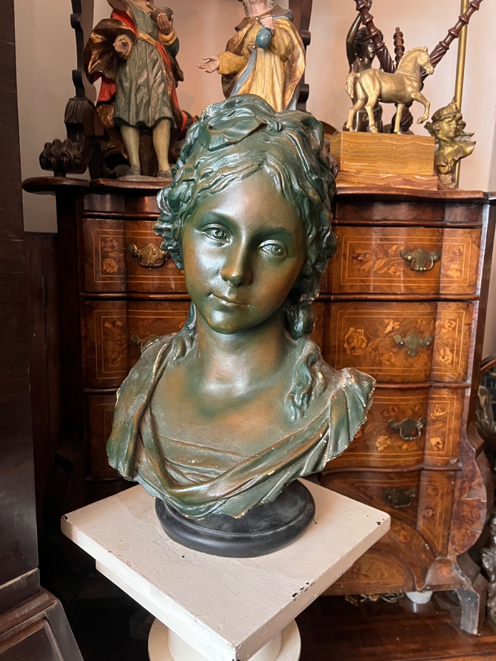 A bronzed terracotta bust of a classical woman featuring a green patinated surface, wavy pinned hair, draped garments, and a dark circular base.