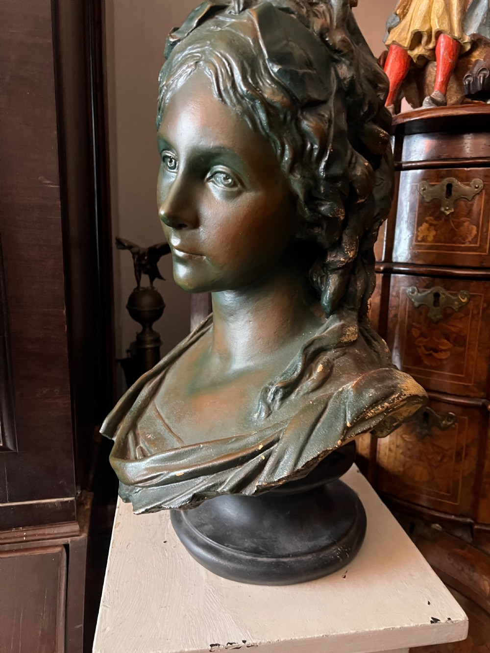 A bronzed terracotta bust of a classical woman featuring a green patinated surface, wavy pinned hair, draped garments, and a dark circular base. View III