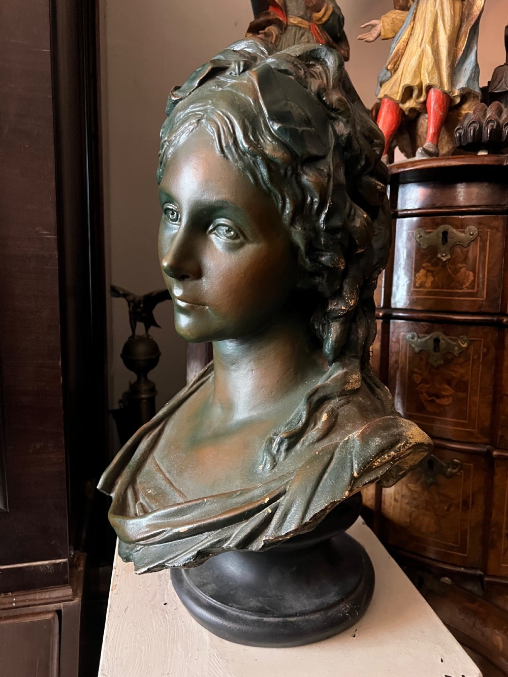 A bronzed terracotta bust of a classical woman featuring a green patinated surface, wavy pinned hair, draped garments, and a dark circular base. View IV