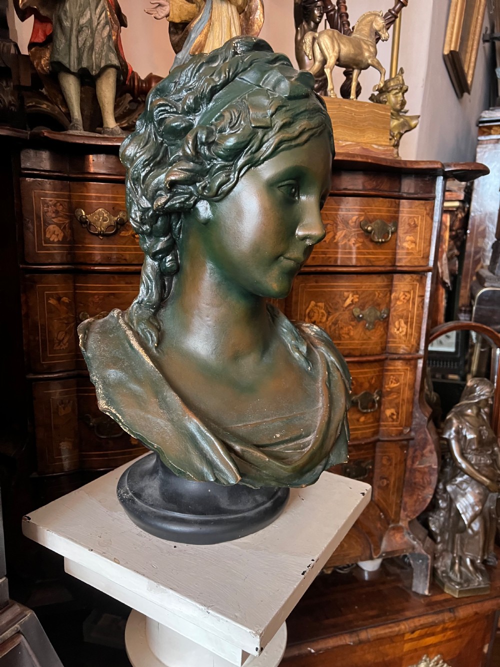 A bronzed terracotta bust of a classical woman featuring a green patinated surface, wavy pinned hair, draped garments, and a dark circular base. View V