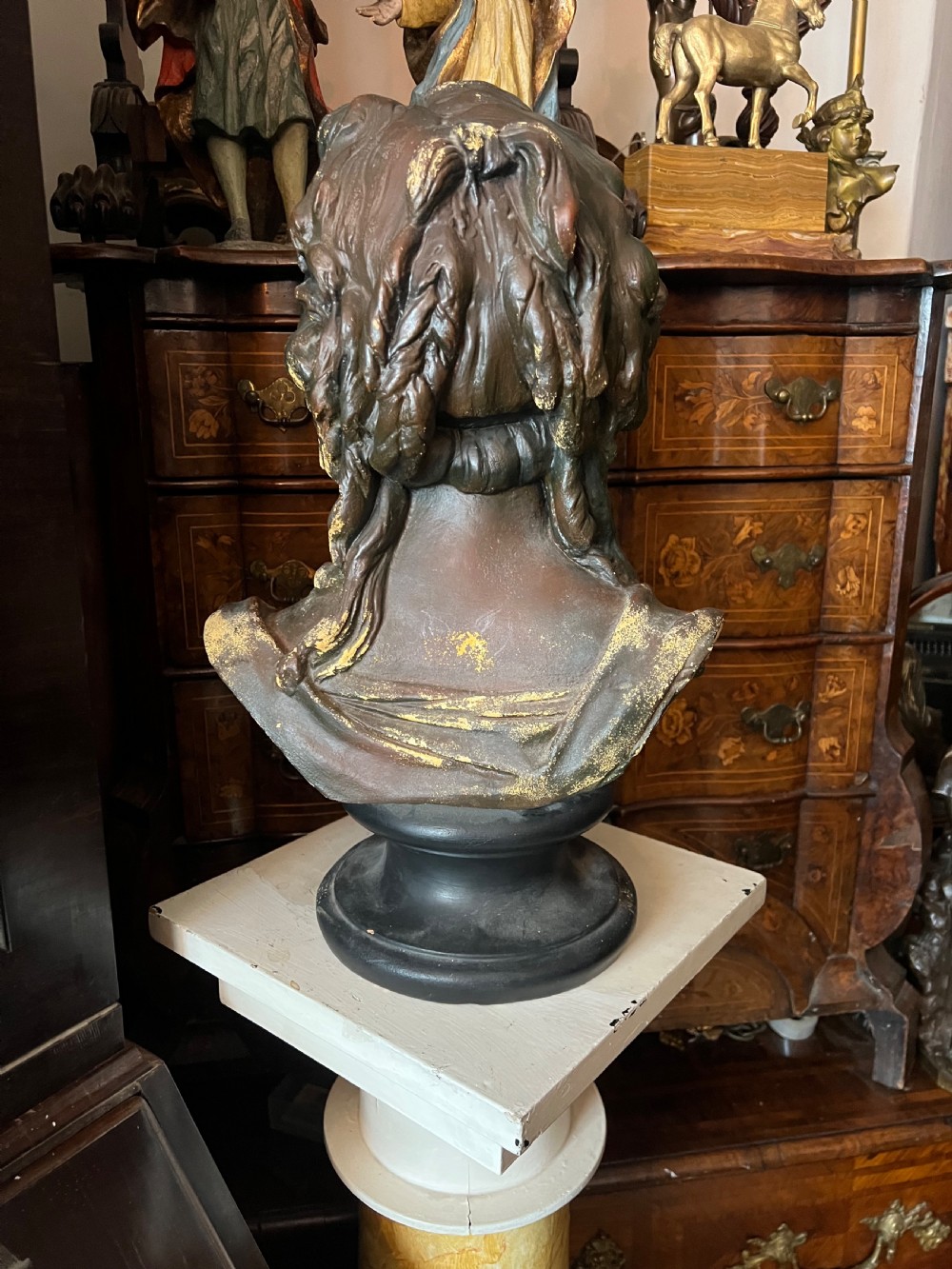 A bronzed terracotta bust of a classical woman featuring a green patinated surface, wavy pinned hair, draped garments, and a dark circular base. View VI