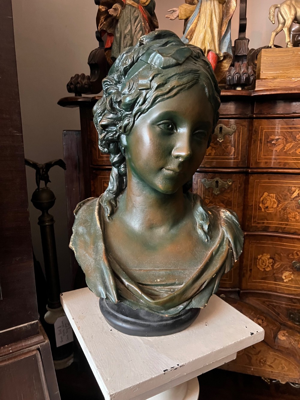 A bronzed terracotta bust of a classical woman featuring a green patinated surface, wavy pinned hair, draped garments, and a dark circular base. View VII