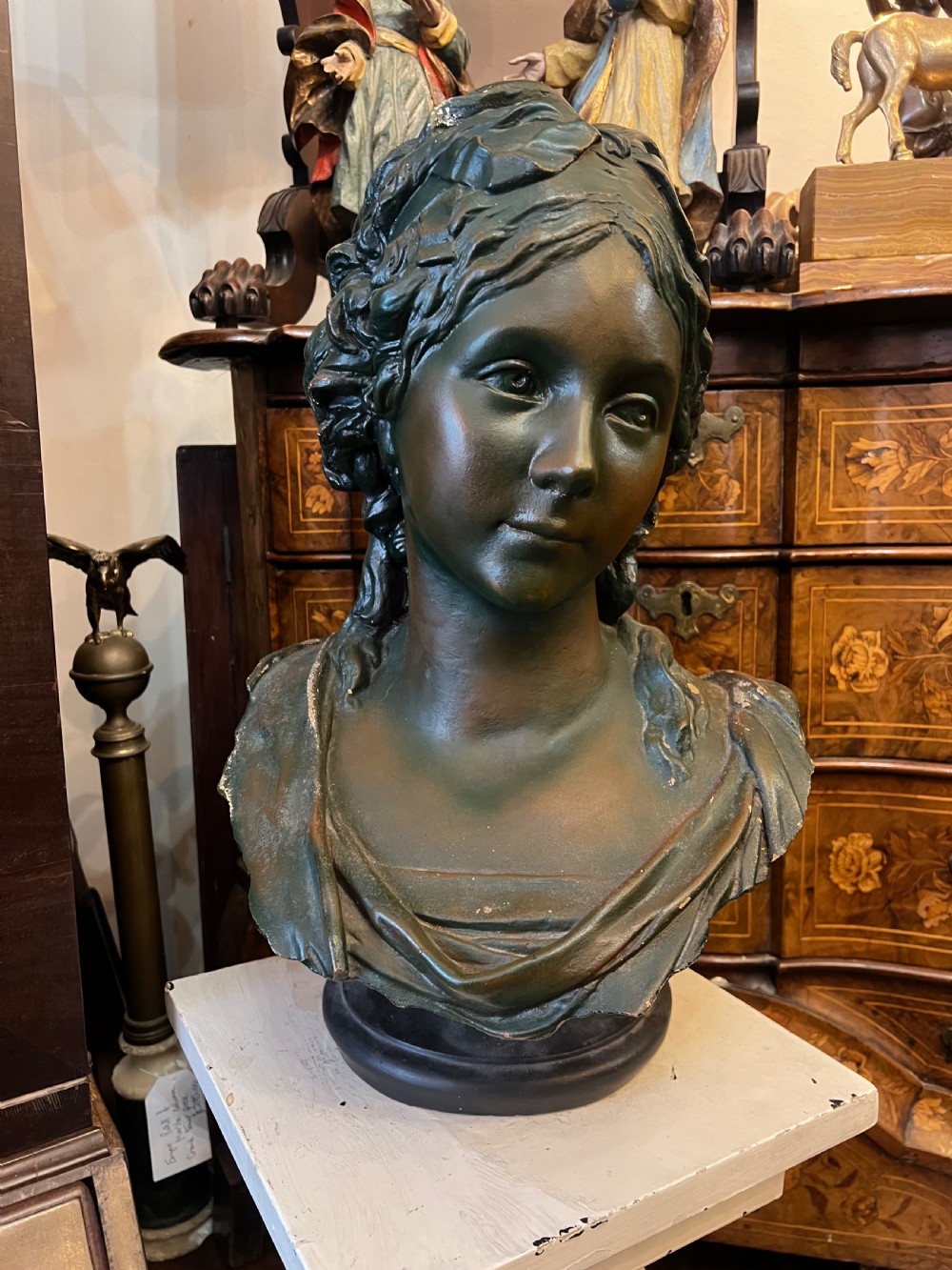 A bronzed terracotta bust of a classical woman featuring a green patinated surface, wavy pinned hair, draped garments, and a dark circular base. View VIII