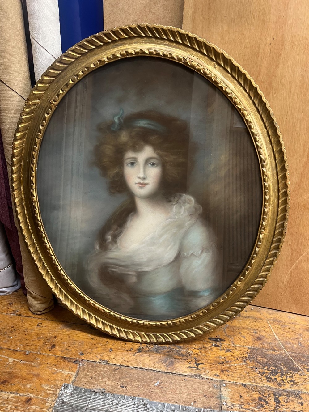 An oval giltwood framed pastel portrait of a young woman with voluminous hair, wearing a powder blue headband and soft cream drape. View II