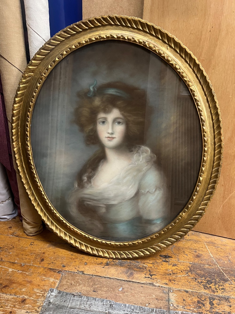 An oval giltwood framed pastel portrait of a young woman with voluminous hair, wearing a powder blue headband and soft cream drape. View IV