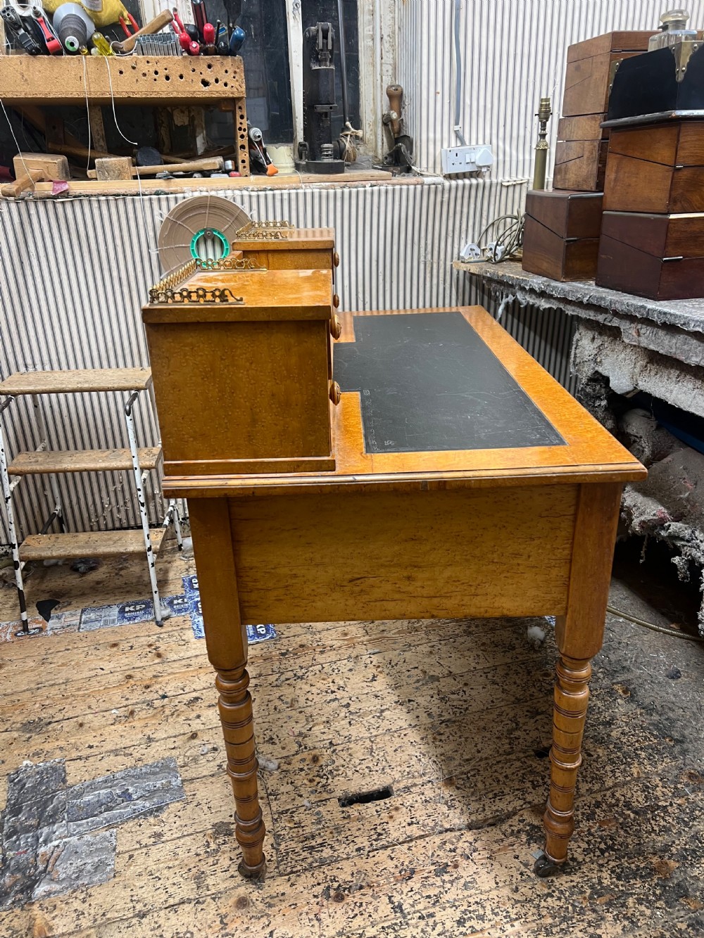 An antique birds eye maple writing desk featuring a black leather surface, turned legs on castors, and an upper superstructure with small drawers and a brass gallery railing. View VI