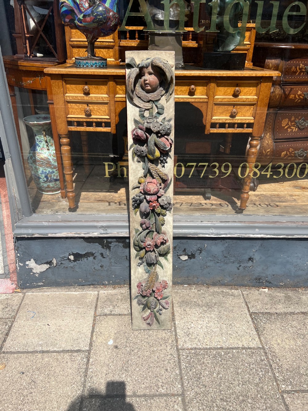 A tall vertical 18th-century carved wooden panel featuring a cherub head above cascading floral and fruit swags painted in muted colours on a light ground. View II