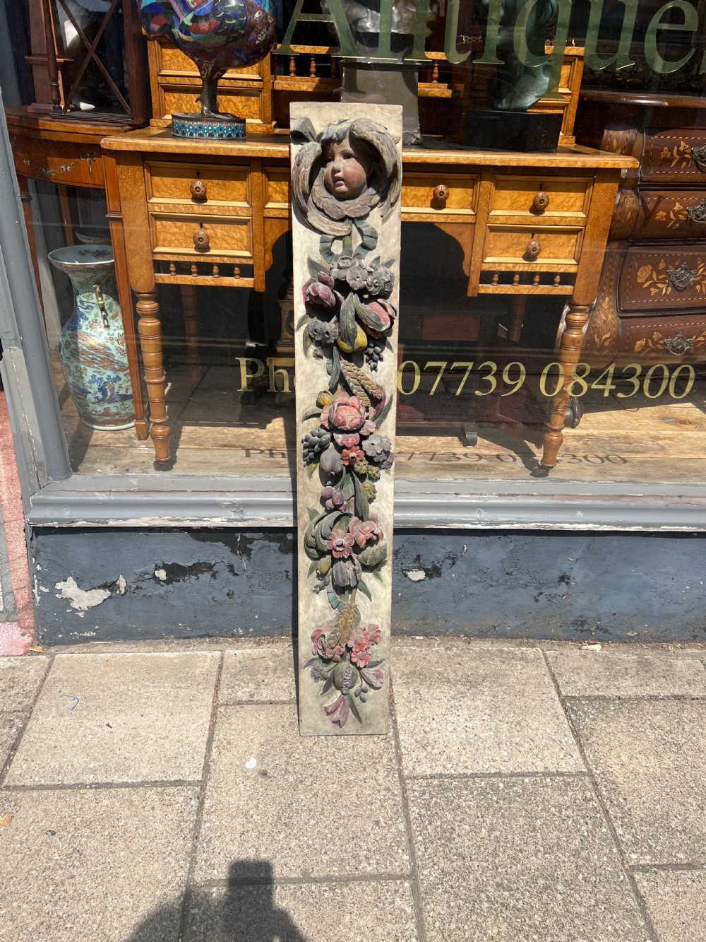 A tall vertical 18th-century carved wooden panel featuring a cherub head above cascading floral and fruit swags painted in muted colours on a light ground. View III
