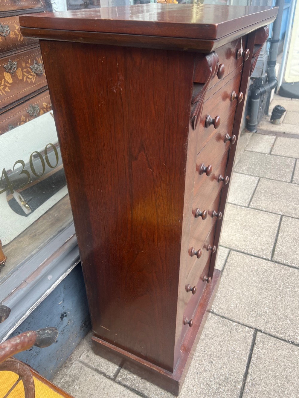 A tall, narrow Victorian walnut Wellington chest featuring seven stacked drawers with wooden turned knobs and carved leaf corbels at the top sides. View III