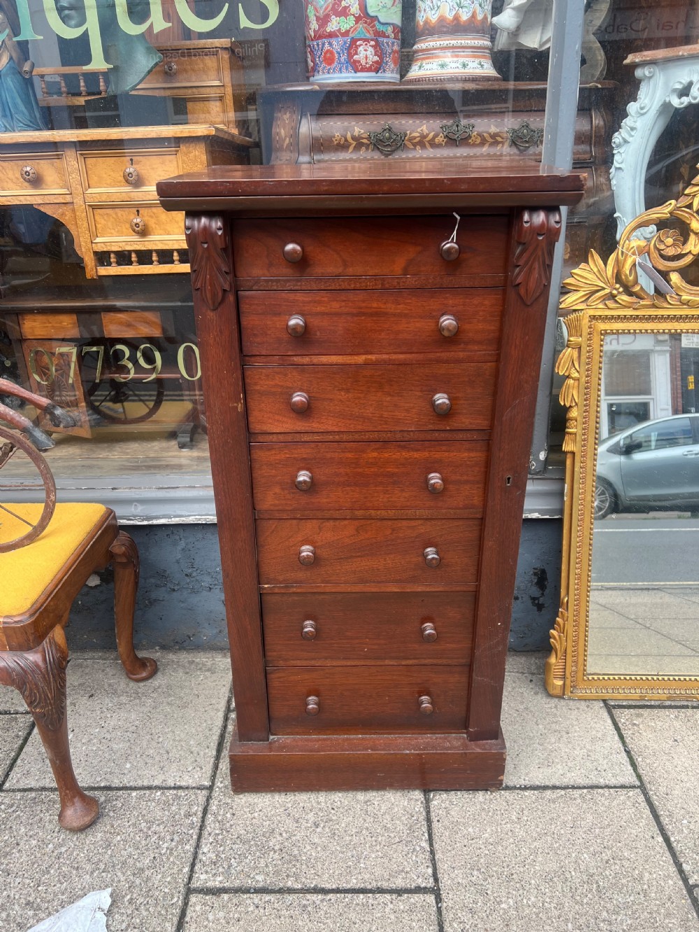 A tall, narrow Victorian walnut Wellington chest featuring seven stacked drawers with wooden turned knobs and carved leaf corbels at the top sides. View VI