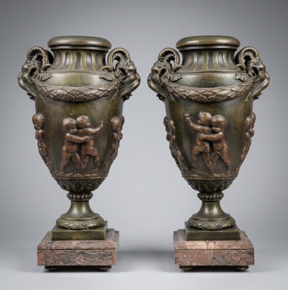 A pair of bronzed spelter Neoclassical urns with relief putti, ram head handles, and square red marble bases.