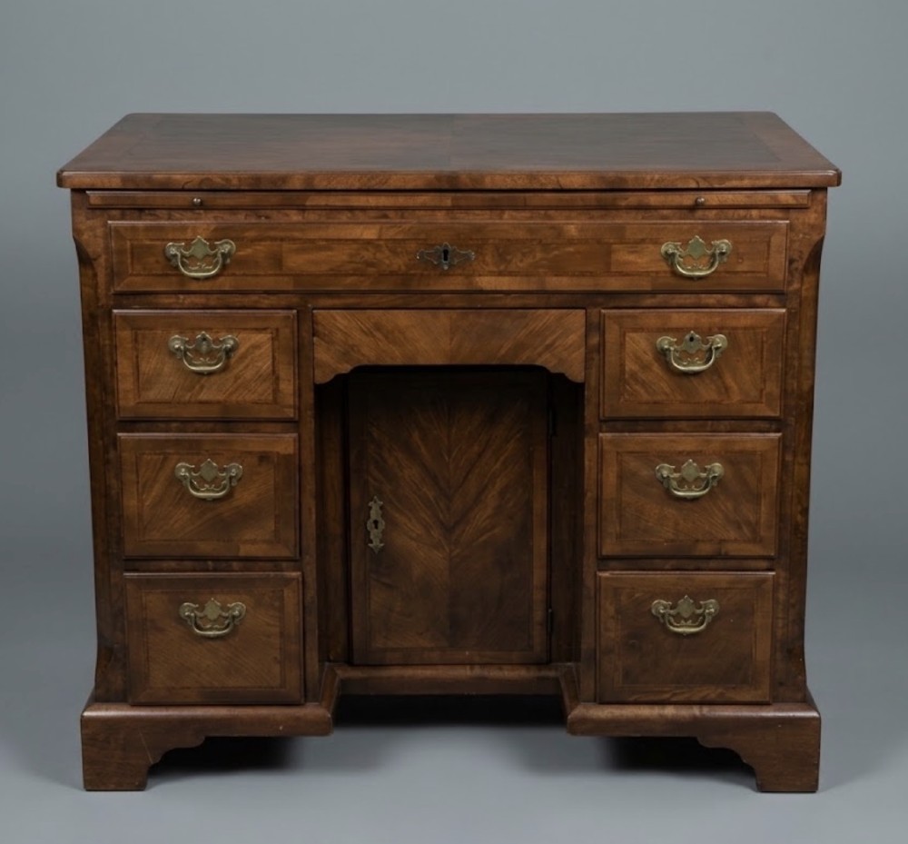 A George II walnut kneehole desk featuring a long top drawer above a recessed central cupboard, flanked by six side drawers with brass swing handles, resting on bracket feet.