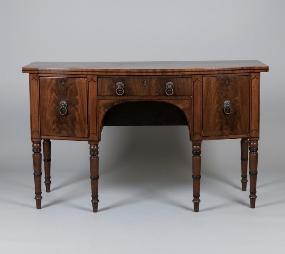 A George III mahogany bowfront sideboard featuring turned legs, lion mask brass handles, central drawer, and flanking cupboards.