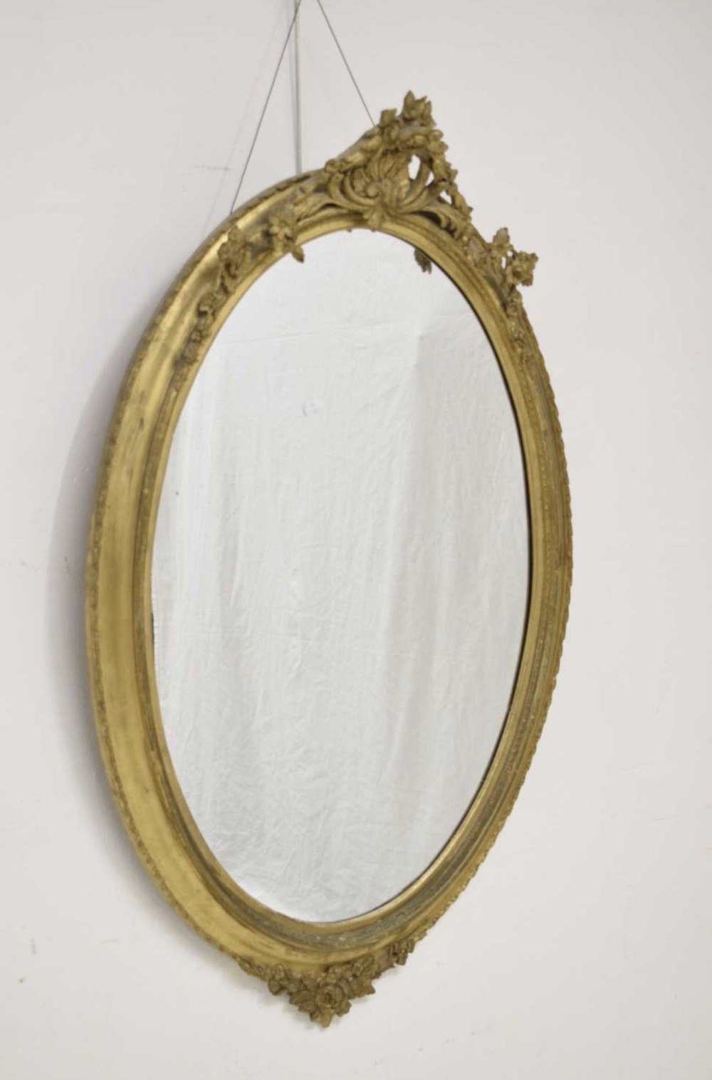 A large oval wall mirror with an ornate gold-gilded wood and gesso frame topped with a carved foliate crest.